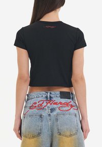 Black cropped t-shirt with short sleeves, featuring red embroidered logo on the back. Paired with blue faded denim shorts and red stitching.