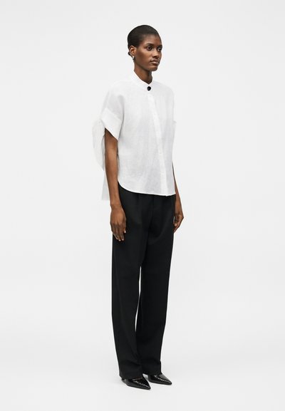 Femme portant une blouse blanche à manches courtes boutonnée, un pantalon noir et des chaussures noires pointues, debout devant un fond blanc uni.