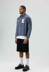 Blauw geruit overhemd met witte knopen en een geborduurde "E" op de borst, gecombineerd met zwarte korte broek en zwarte schoenen met veters. Witte sokken.