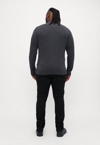 Maglione lungo grigio scuro con maniche lunghe e scollo rotondo, aderente in vita, indossato con pantaloni neri e scarpe, visto da dietro.
