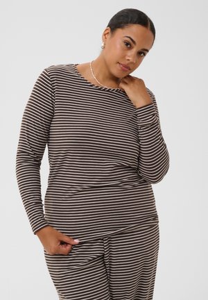 Femme aux cheveux foncés attachés porte un haut à manches longues rayé marron et blanc et un pantalon assorti sur un fond uni.