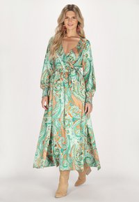 Rochie lungă, verde, cu motive paisley și accente portocalii, decolteu în V, talie strânsă și mâneci ample. Asortată cu cizme cu toc bej.
