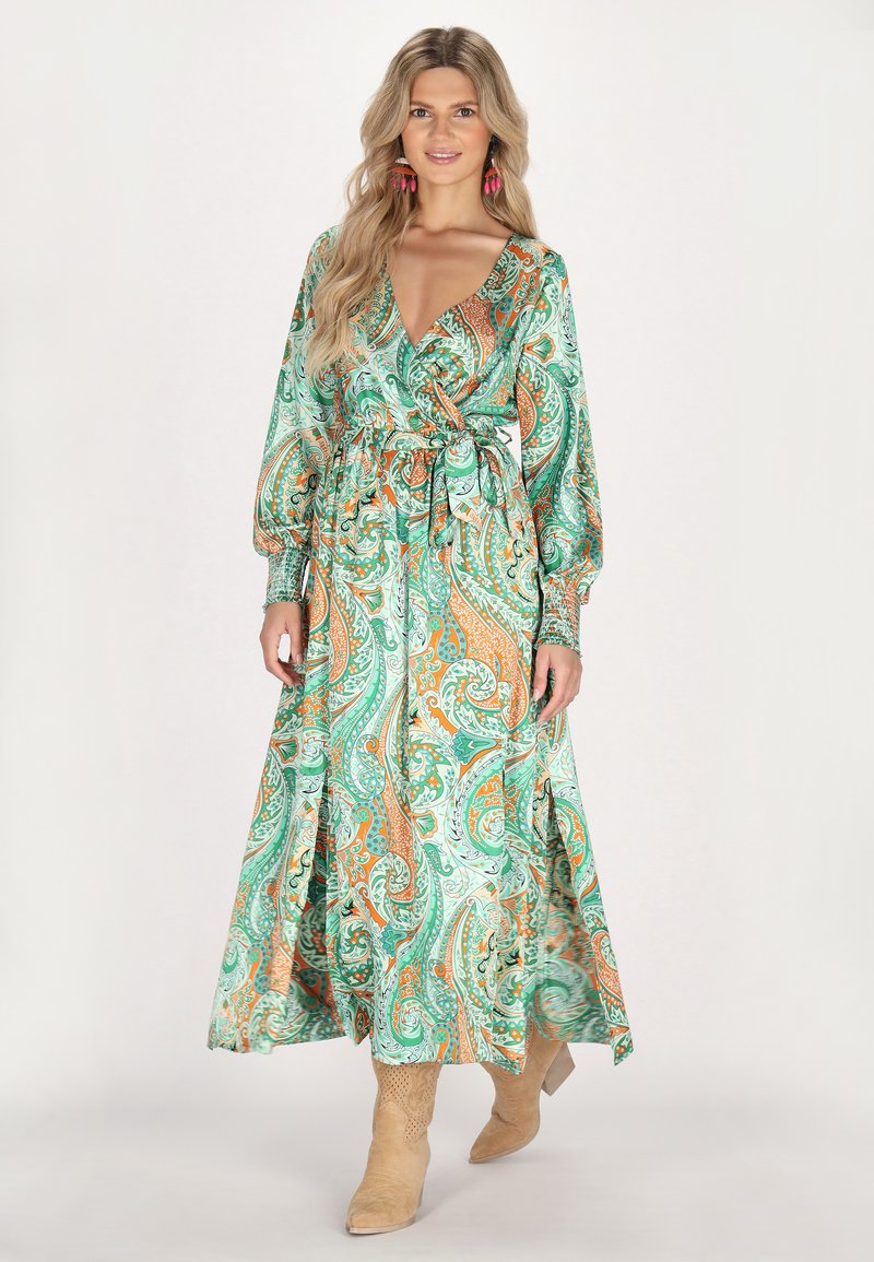 Rochie lungă, verde, cu motive paisley și accente portocalii, decolteu în V, talie strânsă și mâneci ample. Asortată cu cizme cu toc bej.