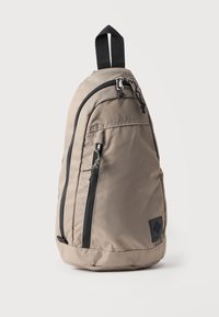 Hellbrauner Sling-Rucksack mit schwarzen Reißverschlüssen, einem Tragegriff oben und einer vertikalen Fronttasche mit Reißverschluss vor weißem Hintergrund.