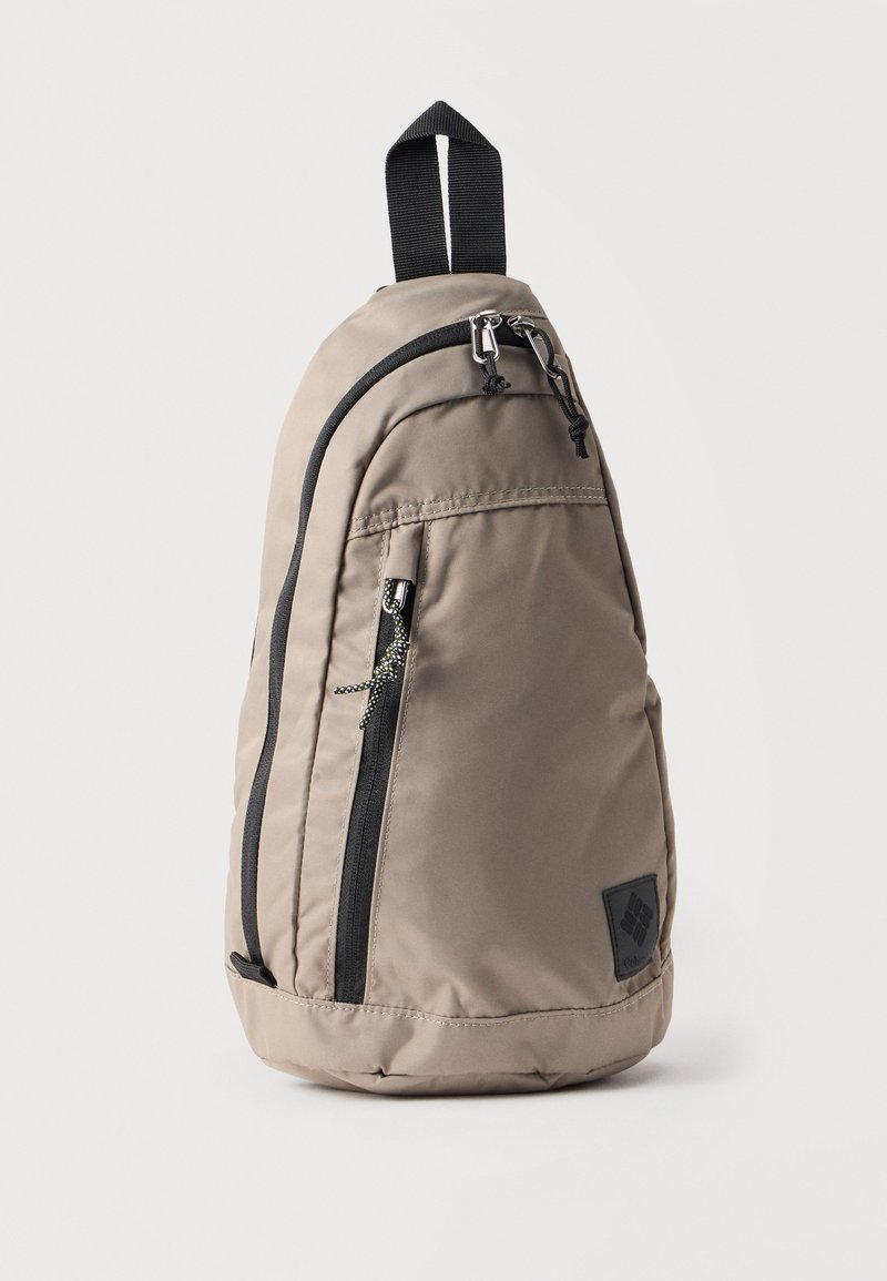 Hellbrauner Sling-Rucksack mit schwarzen Reißverschlüssen, einem Tragegriff oben und einer vertikalen Fronttasche mit Reißverschluss vor weißem Hintergrund.