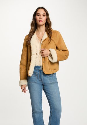 Tanfarbene Wildlederjacke mit Kunstfaser-Schafsleinen-Futter und -Ärmeln, ausgestattet mit zwei Vorderaschen und einem Knopfverschluss. Kombiniert mit hellblauen Jeans.