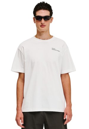 Mann trägt schwarze Sportsonnenbrille, weißes übergroßes T-Shirt mit kleinem schwarzem Text auf der Brust und dunkle Hose, steht vor weißem Hintergrund.