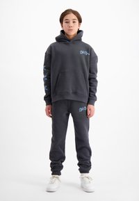 Donkergrijze sweatshirt met capuchon en voorzak, versierd met blauwe borduring. Gepaard met donkergrijze joggers, beide gemaakt van zachte stof. Witte sneakers.