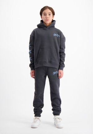 Dunkelgrauer Kapuzenpullover mit Fronttasche und blauer Stickerei. Kombiniert mit dunkelgrauen Jogginghosen, beide aus weichem Material. Weiße Sneakers.