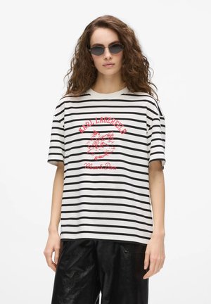 Kvinde med krøllet hår iført mørke solbriller, en sort-hvid stribet T-shirt med rødt katte motiv og sorte vide bukser.