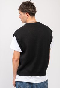 Μαύρος πλεκτός αμάνικος σόρτς με ραβδωτό λαιμό, φορεμένος πάνω από λευκό μπλουζάκι, ματιά από πίσω, επιδεικνύοντας χαλαρή εφαρμογή.