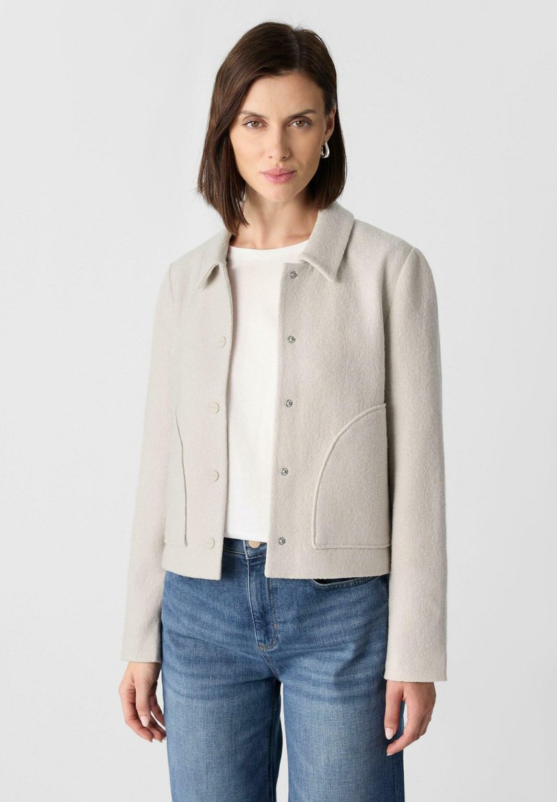 Hellgraue, kurze Jacke aus strukturiertem Stoff mit Druckknöpfen, spitzem Kragen und seitlichen Taschen. Getragen über einem weißen Hemd und blauen Jeans.
