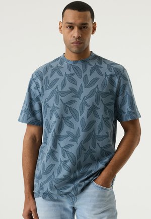 Man die een blauw shirt met korte mouwen en donkere bladpatronen draagt, staat met één hand in de zak van een lichtblauwe spijkerbroek.