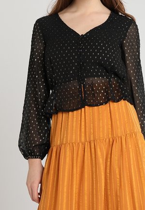 Kobieta w czarnej, przezroczystej bluzce z długim rękawem, ozdobionej złotymi akcentami i zapinanej na guziki, w połączeniu z pomarańczową, plisowaną spódnicą z wysokim stanem.