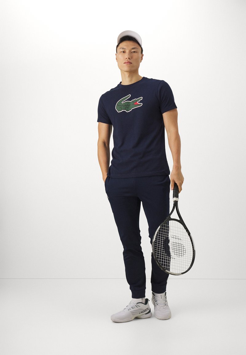 Maglietta blu navy con un logo di coccodrillo verde e rosso, abbinata a pantaloni navy e sneakers bianche, mentre tiene una racchetta da tennis.