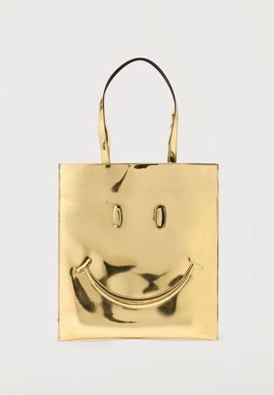 Kuldse metallilise välimusega käekott pikkade sangadega, mille esiküljel on reljeefne naeratava näo kujundus ühtlase taustaga.