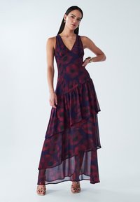 Maxi dress in sheer dark purple fabric with red floral pattern, v-neckline, tiered ruffles, and ankle-length hem. Worn with gold accessories.