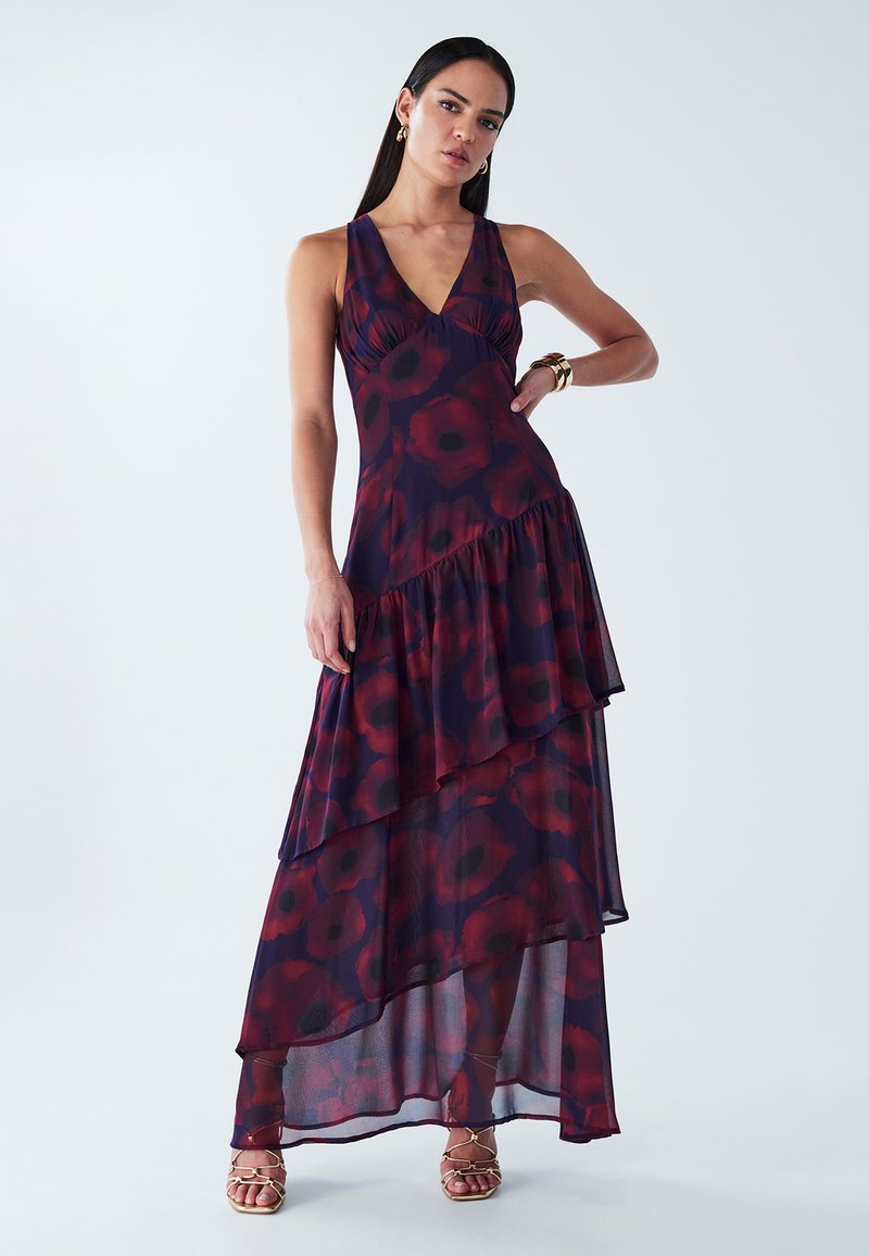 Maxi dress in sheer dark purple fabric with red floral pattern, v-neckline, tiered ruffles, and ankle-length hem. Worn with gold accessories.