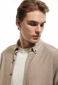 Camisa de botones a rayas beige con botones marrones, llevada sobre una camiseta de color claro. El cabello está peinado de manera casual y lleva un aro de pendiente.