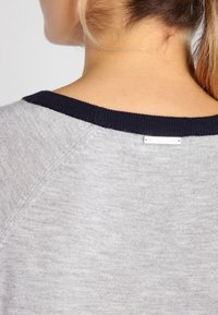 Jersey gris con cuello acanalado en azul marino, presenta una textura suave y una costura elegante. Etiqueta visible en la parte trasera, que muestra el nombre de la marca.