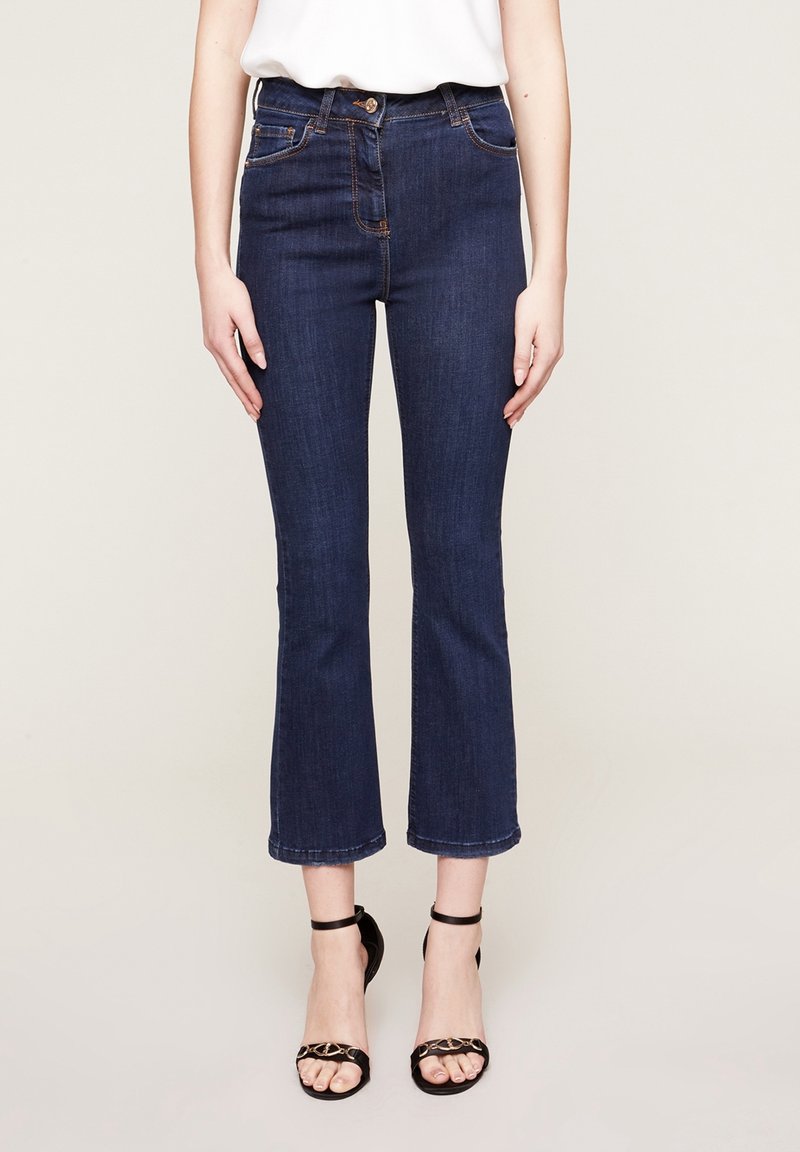 Femme portant un jean flare court bleu foncé avec bouton et fermeture éclair, associé à des talons noirs ouverts à bride cheville et un haut blanc.