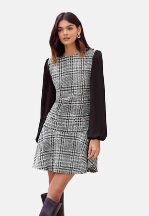 Femme portant une robe à carreaux noire et blanche avec de longues manches bouffantes noires et des bottes noires montant jusqu'aux genoux, debout devant un fond uni.