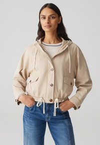 Giacca beige cropped con cappuccio, chiusura a bottoni, vita con cordino e due tasche frontali. Abbinata a jeans blu. Texture liscia.