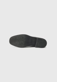 Semelle de chaussure noire en caoutchouc texturé, présentant un motif en zigzag et un logo de la marque embossé dans la zone du talon. Forme plate et légèrement courbée.