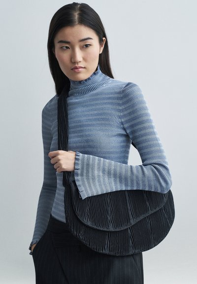 Giovane donna con un dolcevita blu a fantasia che tiene in mano una grande borsa a spalla nera plissettata, mentre guarda di lato su uno sfondo neutro.