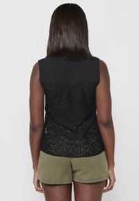 Top neagră fără mâneci, cu un voal solid în față și un spate din dantelă complicată. Combinată cu pantaloni scurți de culoare verde măsliniu, oferind un aspect relaxat și un stil casual.