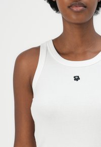Hvit ribbestrikket tanktopp med rund hals og svart blomsterbroderi på brystet. Stoffet ser mykt og stretchy ut.