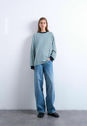 Jeune femme debout contre un mur blanc portant une chemise à manches longues rayée bleu et blanc, un jean bleu à jambes larges et des chaussures noires.