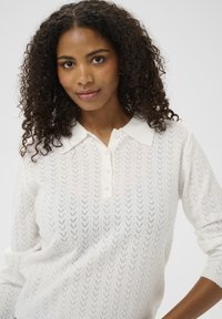 Pull en tricot blanc avec un motif en V, doté d'un col et de quatre boutons. Le tissu est léger et texturé.