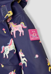Impermeable azul marino con coloridos unicornios y patrones florales, con un parche rectangular rosa que dice "JoJo splish splash" en la manga.