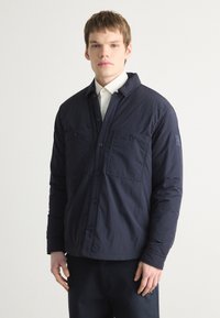 Chaqueta de camisa ligera en azul marino con cierre de botones, dos bolsillos frontales y un diseño moderno que presenta un tejido texturizado y un ajuste relajado.