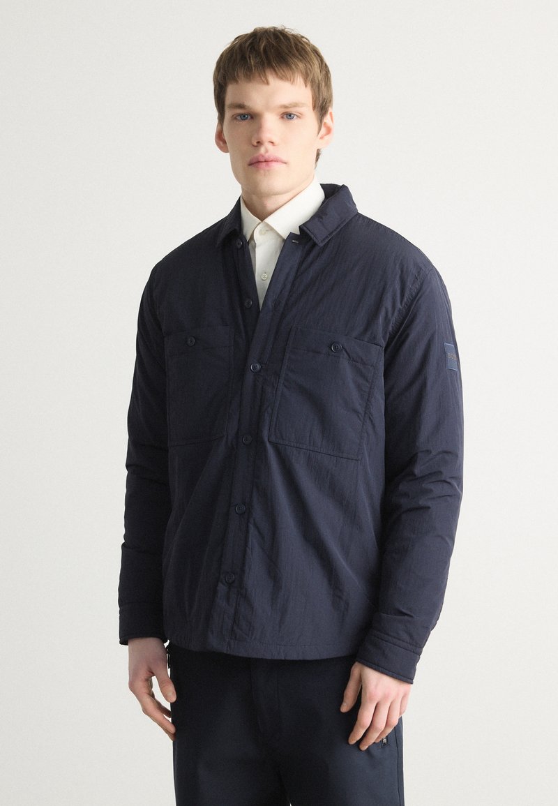 Chaqueta de camisa ligera en azul marino con cierre de botones, dos bolsillos frontales y un diseño moderno que presenta un tejido texturizado y un ajuste relajado.