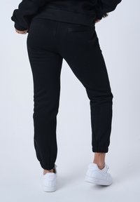 Pantalones jogger negros hechos de una mezcla de algodón, con un diseño ajustado, puños elásticos y un bolsillo trasero. Llevados con zapatillas blancas.