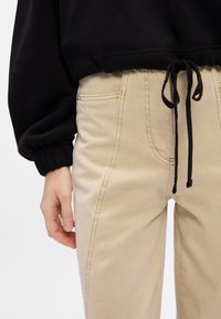 Pantaloni beige a vita alta con finitura testurizzata, dotati di tasche laterali e dettagli in cordino nero in vita, abbinati a un maglione nero.