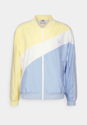 Homme portant une veste pastel jaune, blanche et bleue, un t-shirt blanc, un short noir, des chaussettes blanches et des baskets Adidas vertes, debout devant un fond uni.