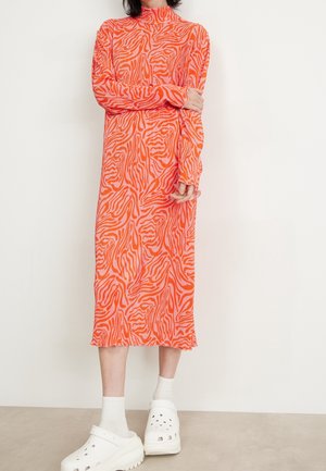 Osoba ubrana w długą sukienkę midi w pomarańczowo-różowy wzór zebry, białe skarpetki i masywne białe chodaki, stojąca na tle jednolitej ściany.