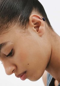 Pendiente de aro de plata incrustado con una piedra azul oscura, detallado con pequeños acentos negros, exhibido en una oreja, con cabello negro liso.