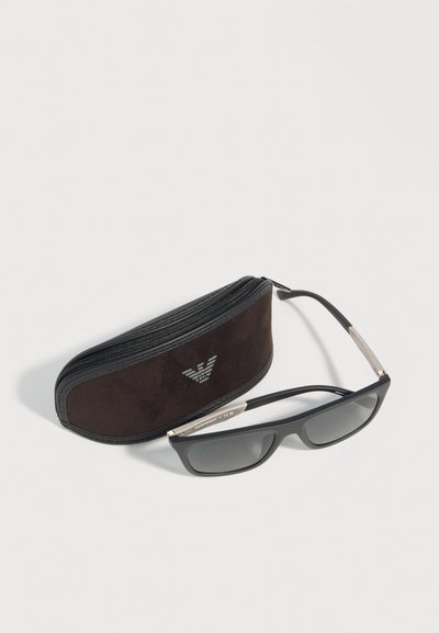Μαύρα ορθογώνια γυαλιά ηλίου με ασημένιες λεπτομέρειες στα μπράτσα, που ακουμπούν δίπλα σε μια μαύρη σκληρή θήκη με φερμουάρ και λευκό λογότυπο.