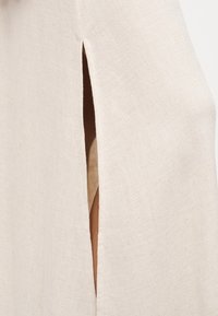 Μπεζ λινοΰ ύφασμα με μια κάθετη σχισμή που αποκαλύπτει ένα μικρό τμήμα δέρματος από κάτω, δείχνοντας υφή και λεπτομέρειες φωτεινής σκιάς.