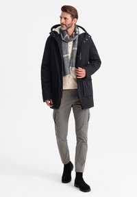 Parka nera isolata con cappuccio, foderato in crema, sciarpa grigia con motivo a spina di pesce, maglione beige chiaro, pantaloni cargo grigi e stivali neri.