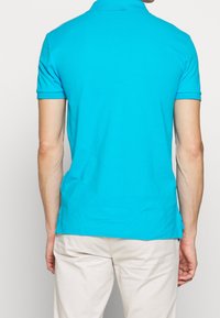 Tyrkysová polo košile s krátkými rukávy a klasickým límcem, vyrobená z hladké látky, s rovým lemem a bez viditelných vzorů či akcentů.