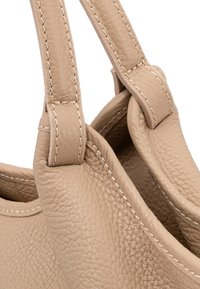 Beżowa skórzana torebka z teksturowaną powierzchnią, z przeszywanymi uchwytami i zaokrąglonymi krawędziami, co zapewnia spójny design. Brak widocznych elementów metalowych.