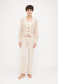 Beige knitted cardigan with three buttons, worn over a matching top and flared pants, all in soft, lightweight material.