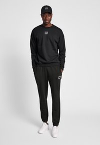 Man med svart keps, svart sweatshirt, svarta joggers med vita logotyper och vita sneakers, stående med händerna i fickorna mot en grå bakgrund.