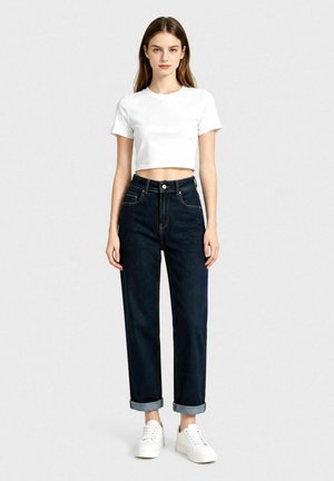 Giovane donna in piedi, indossa una t-shirt bianca corta, jeans scuri risvoltati e sneakers bianche, su uno sfondo chiaro e semplice.