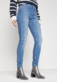 Jeans ajustados de color azul claro hechos de denim, con cintura alta, diseño de cinco bolsillos, costuras visibles y combinados con botas de tacón metálicas hasta el tobillo.